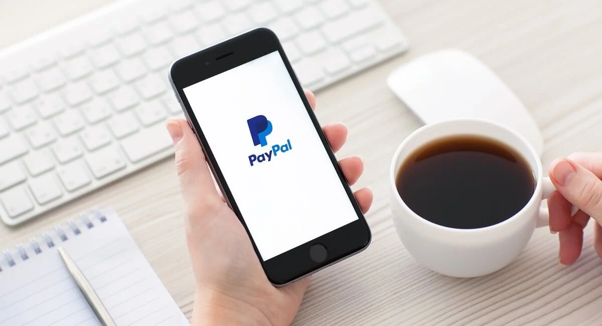 Mão segurando celular com logo PayPal na tela e outra segurando xícara de café