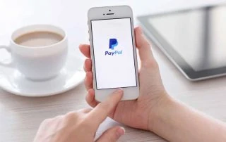 Maõs segurando celular com logomarca da PayPal com xícara de café e tablet ao fundo