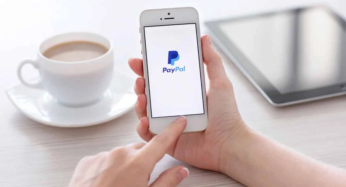 Maõs segurando celular com logomarca da PayPal com xícara de café e tablet ao fundo