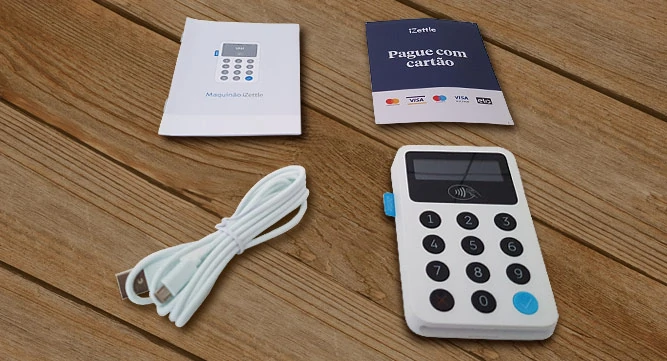 Maquinão iZettle sobre mesa ao lado de cabo USB, manual de instruções e adesivos de bandeiras