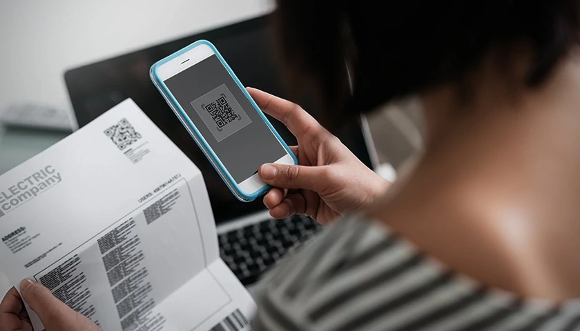 Ilustração de uma mulher realizando pagamentos com celular por QR Code, com um laptop de fundo