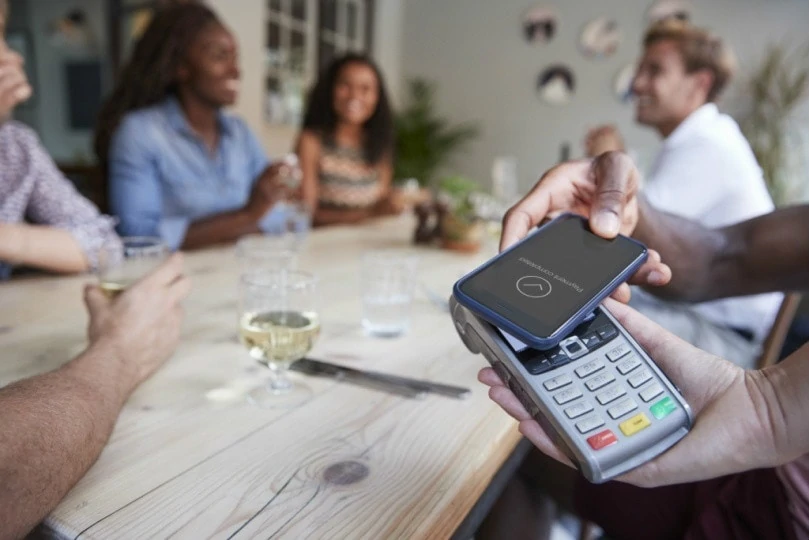 Ilustração mostrando pagamento sem contato com entre celular e máquina de cartão, com pessoas sentadas em uma mesa