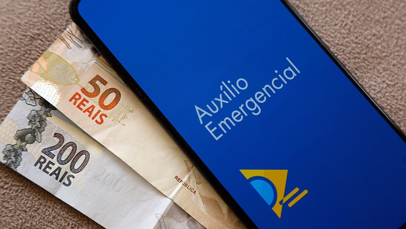 Logo do Auxílio Emergencial na tela de celular ao lado de cédulas de reais